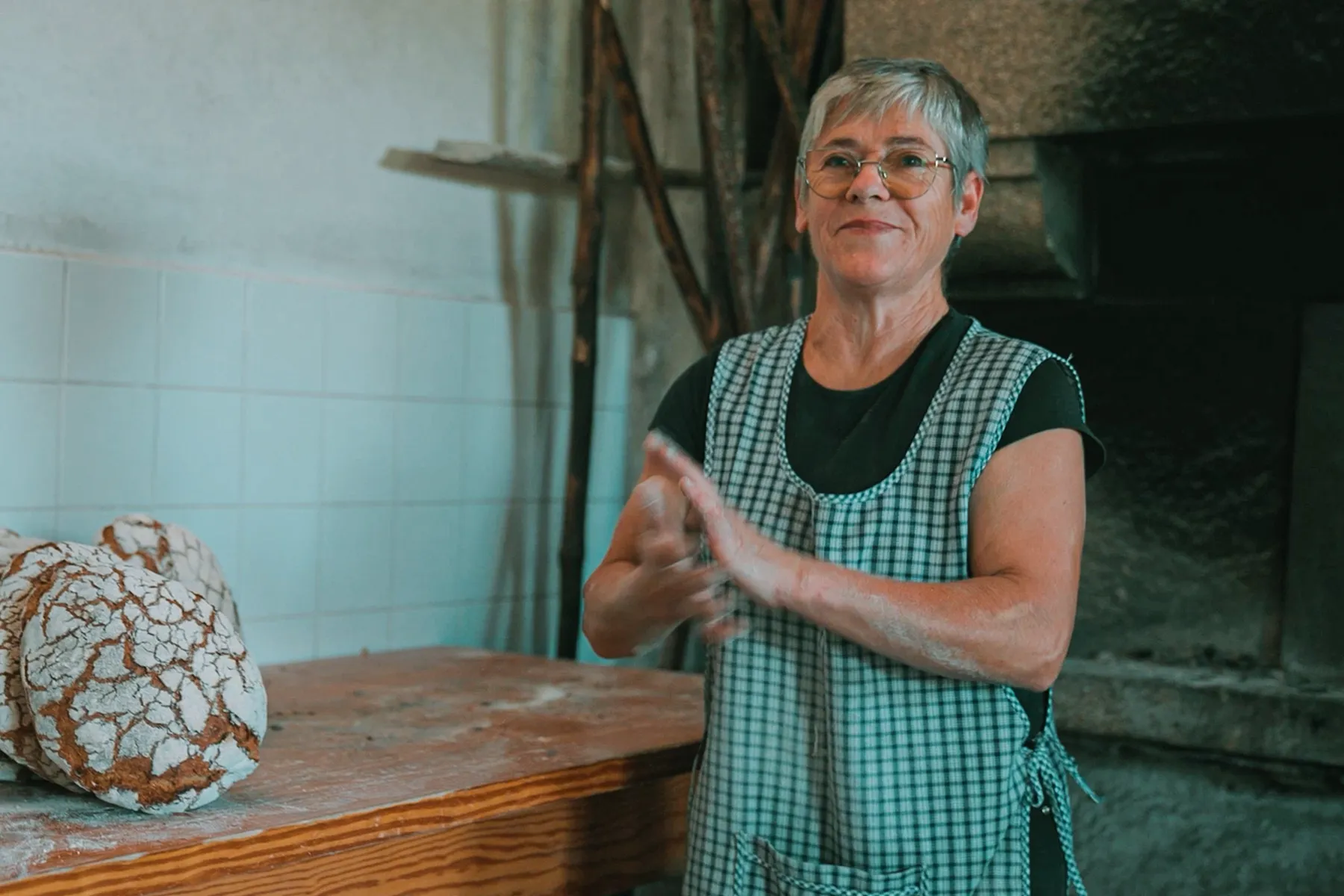 Padeira sorrindo em frente a forno tradicional de pães rústicos.